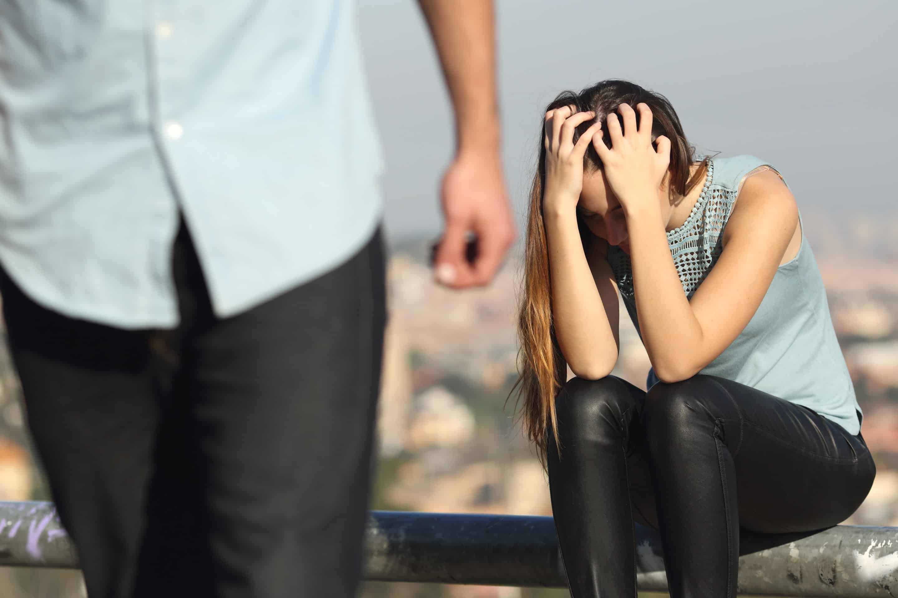 Femme assise, en détresse, tenant sa tête entre les mains tandis qu'un homme s'éloigne en arrière-plan, symbolisant la jalousie destructrice d'un pervers narcissique.