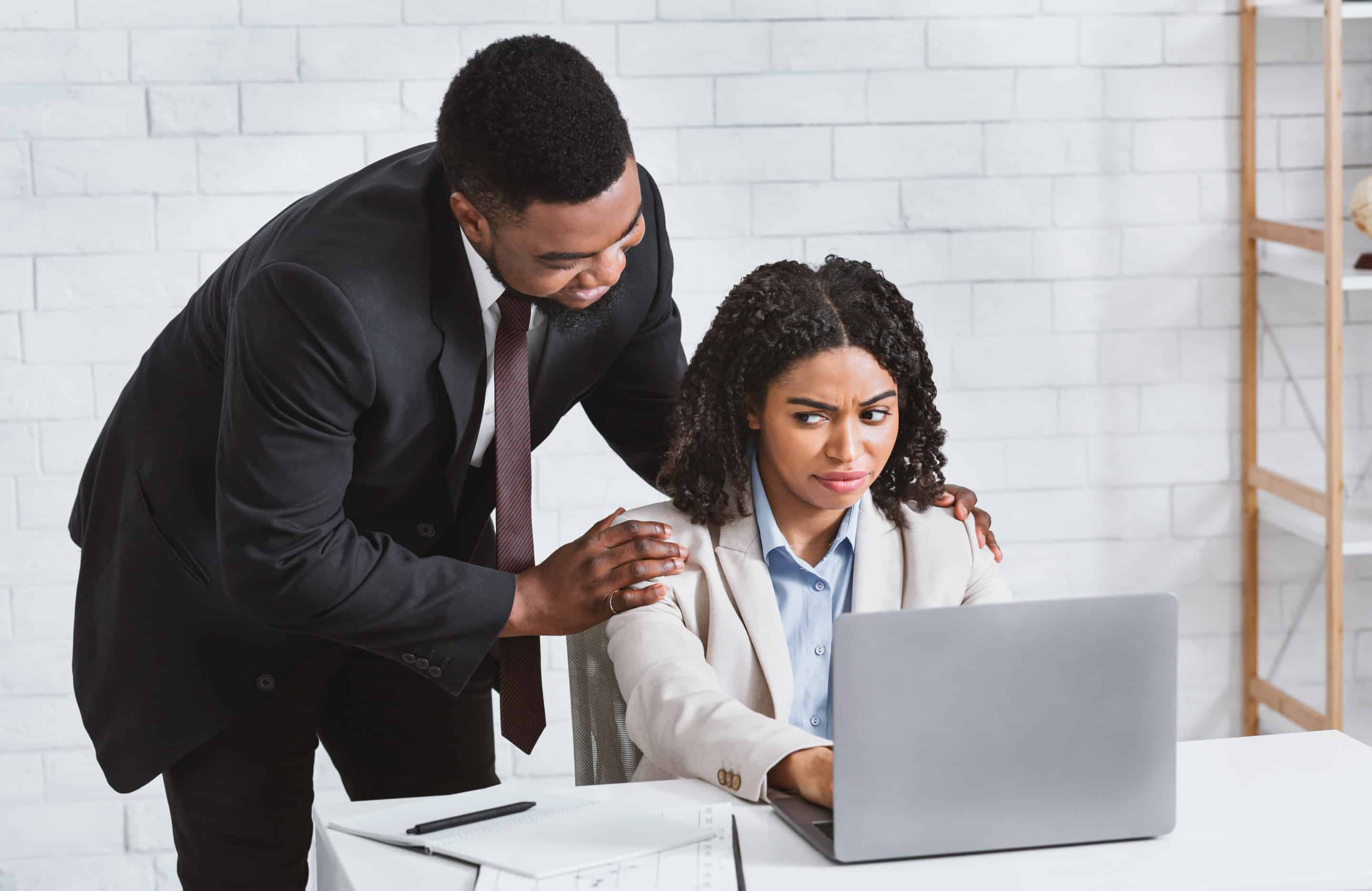 Un homme en costume pose ses mains sur les épaules d'une femme visiblement mal à l'aise, assise devant un ordinateur, symbolisant le stress et l'inconfort au travail, souvent associés au burnout.