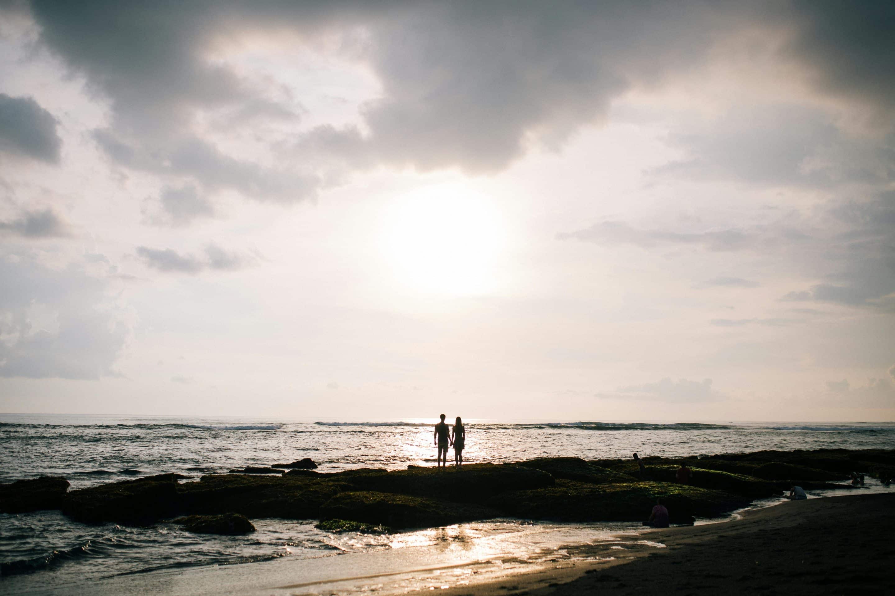Silhouette d’un couple debout sur des rochers au bord de l’océan, au coucher du soleil. L’image évoque à la fois la connexion intime et l’isolement, illustrant la dualité entre passion et emprise dans une relation toxique.